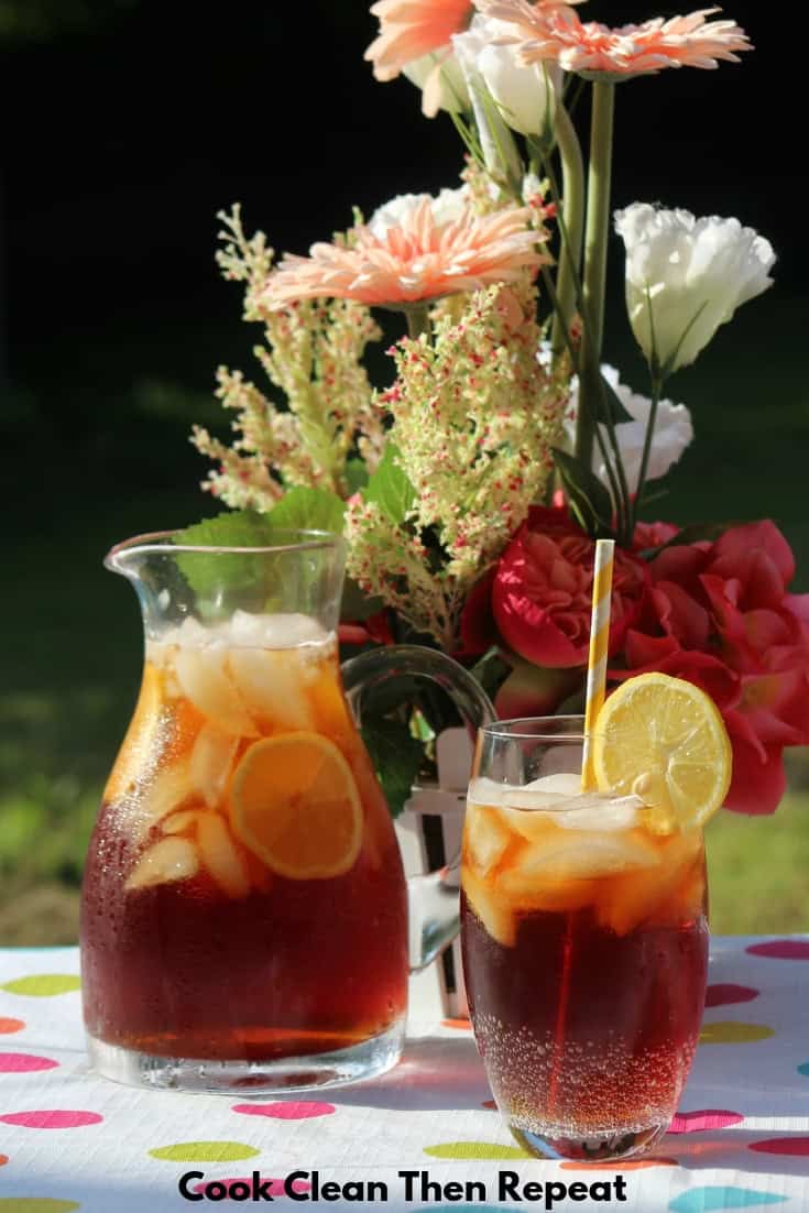 Sweet tea in a pitcher on a table with a glass next to it with some sweet tea and ice in it with a lemon slice and a yellow and white paper straw. Flowers in the background.