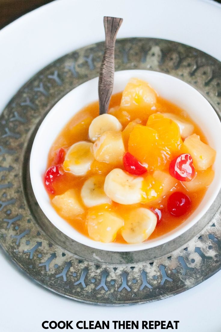 image of the fruit salad in a tall image. fruit salad in a white bowl with spoon on it and plate under the bowl is also silver. 