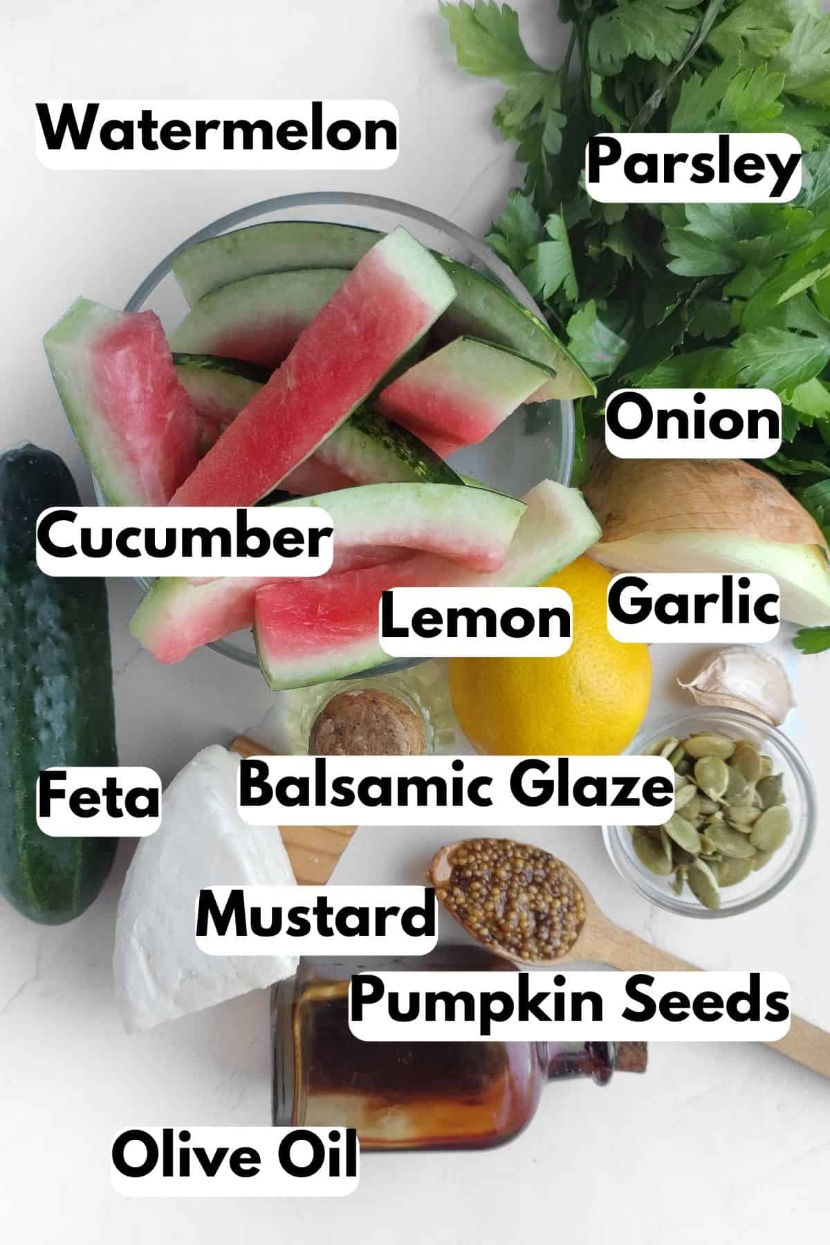 Ingredients needed for watermelon feta salad: watermelon, parsley, onion, cucumber, lemon, garlic, feta, balsamic glaze, mustard, pumpkin seeds, and olive oil.