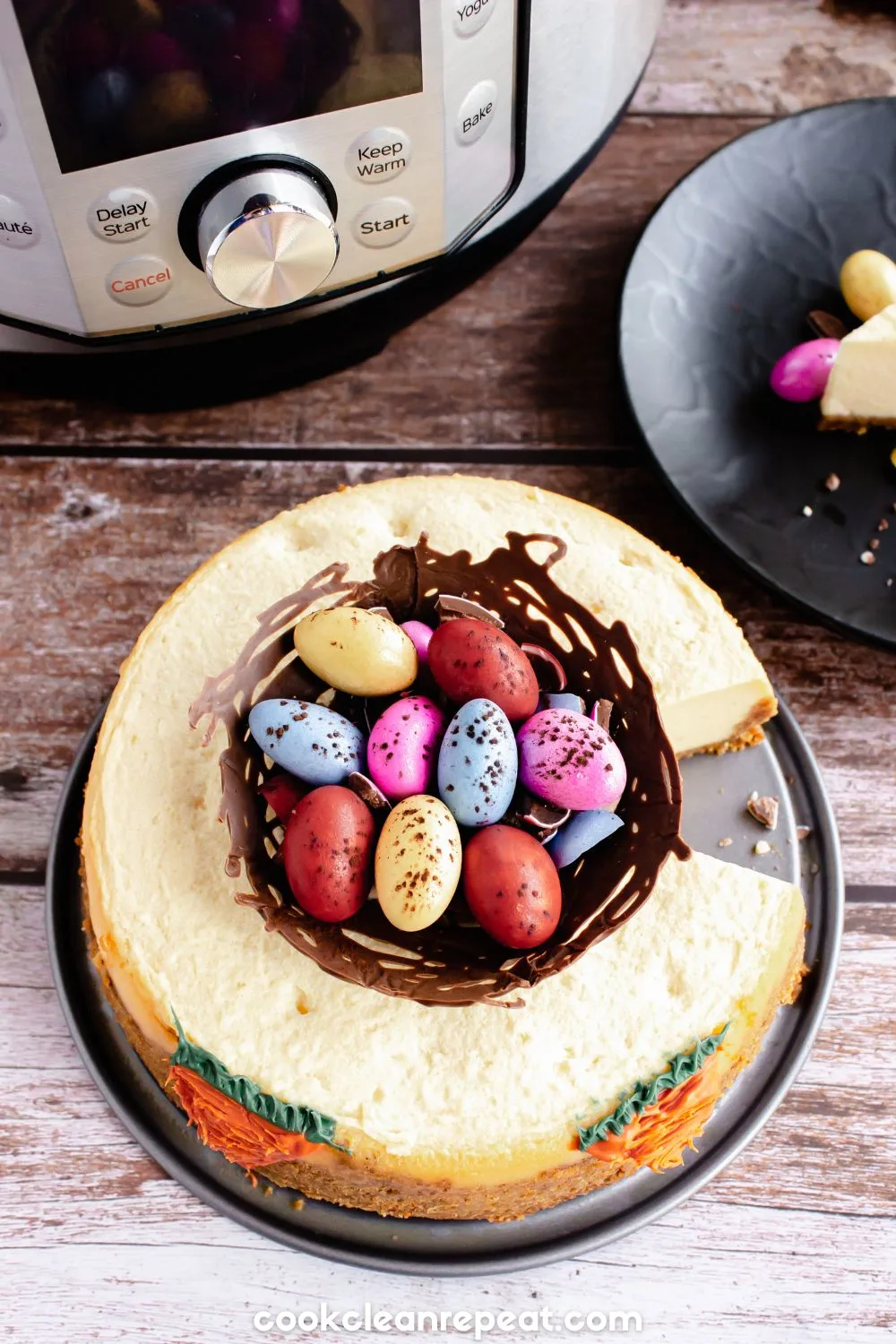 Easter cheesecake recipe completed with a chocolate nest filled with chocolate eggs on top. There are carrots piped onto the side of the cake.