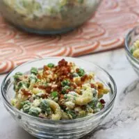a bowl of pea and pasta salad on a marble background with a dish towel and spoon and a bowl filled with the salad in the background