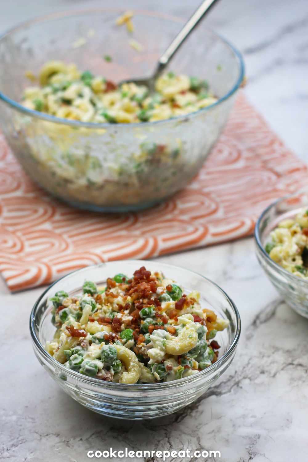a bowl of pea and pasta salad on a marble background with a dish towel and spoon and a bowl filled with the salad in the background