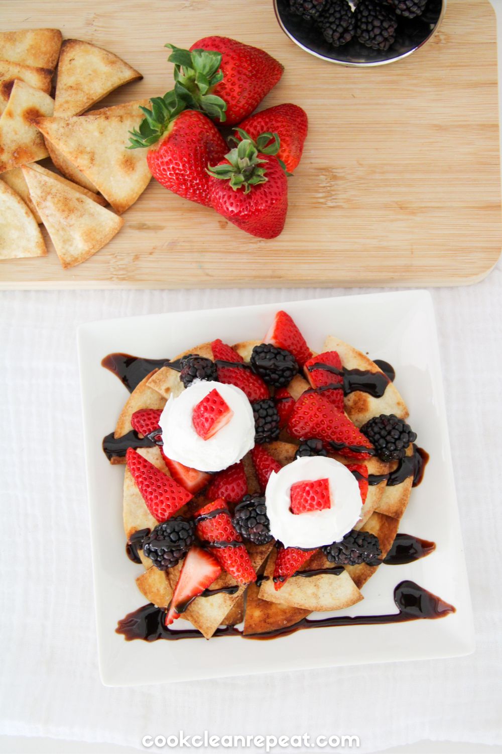 dessert nachos served on a white plate topped with whipped cream and strawberry pieces. There is a cutting board with other chips and strawberries above the plate.