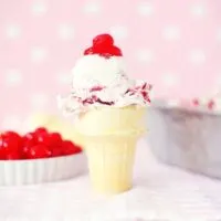 cherry cheesecake ice cream scooped into a cone with cherries on top in front of a pink background